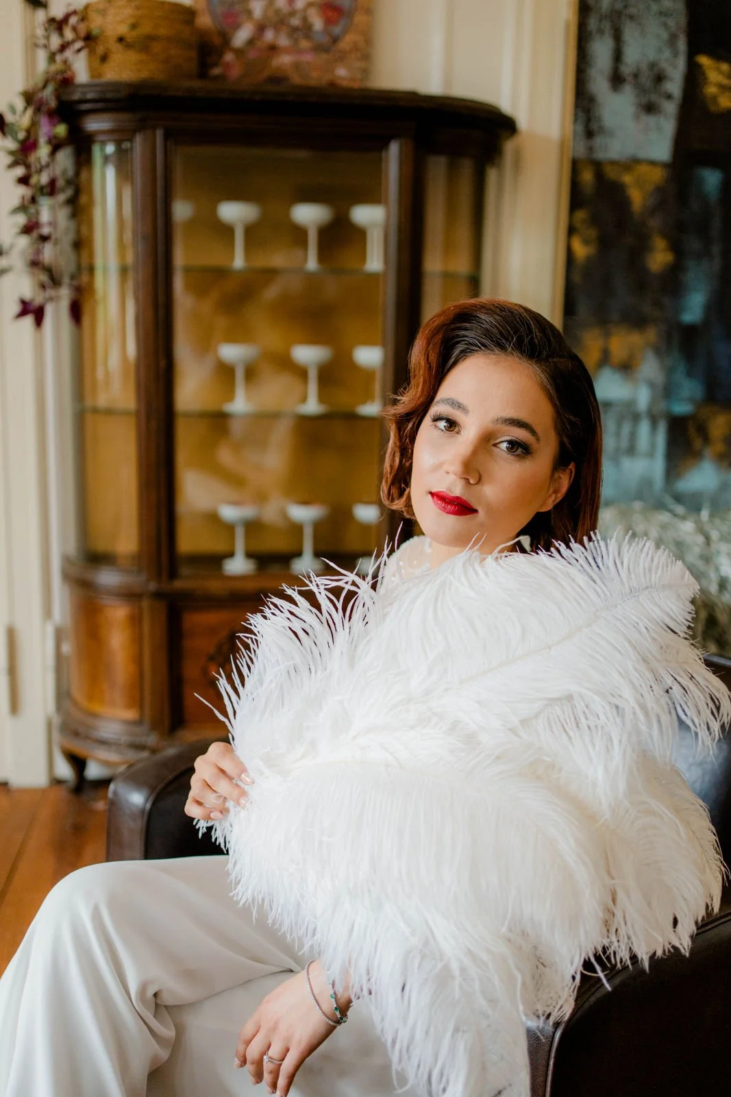Eine Frau mit rotem Lippenstift und braunen Haaren, die ein weißes Federkleid trägt, sitzt auf einem schwarzen Sessel vor einem Holzschrank mit weißen Dessert-Gästallern.