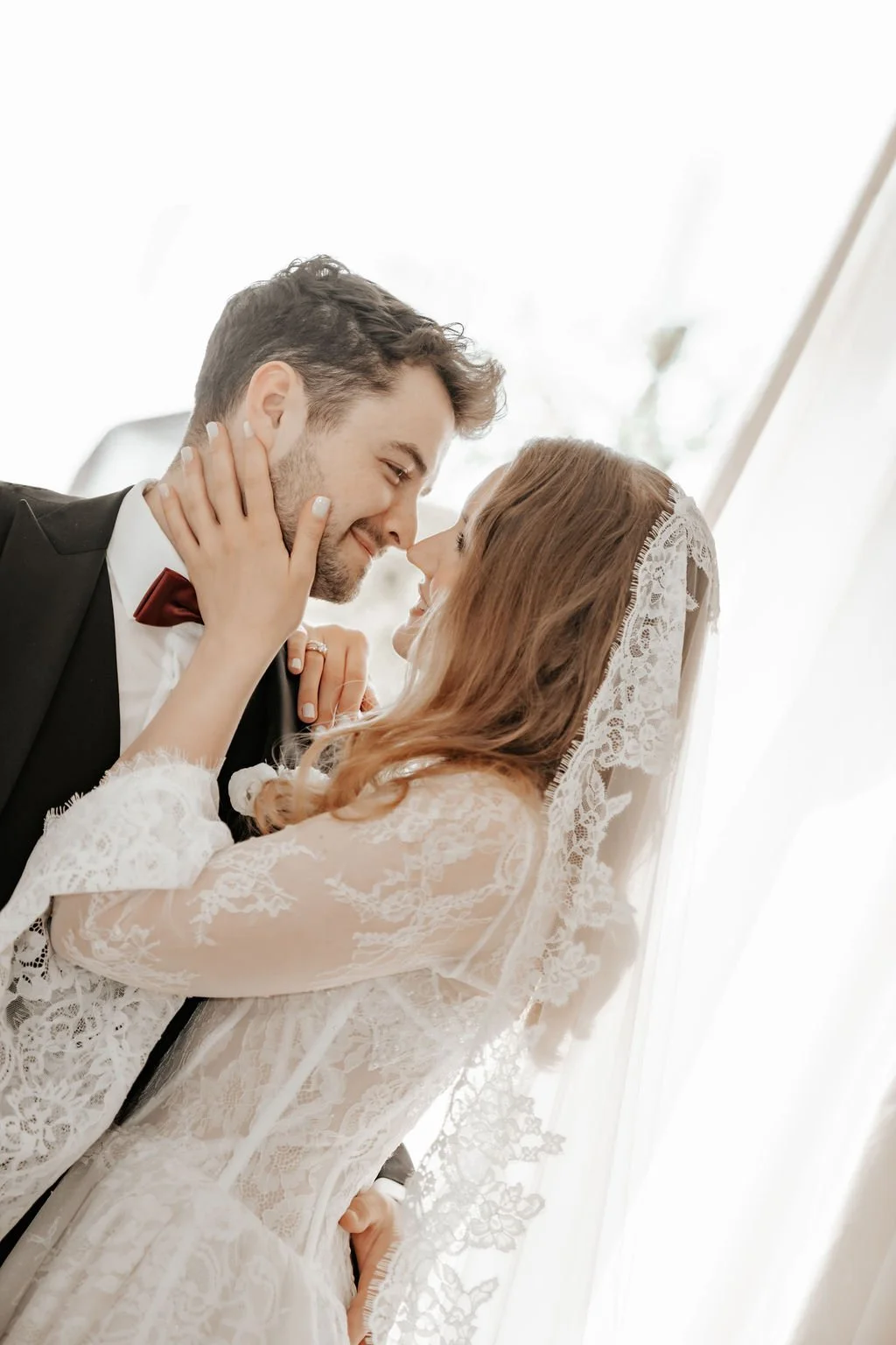 Ein Brautpaar steht sich nahe und lächelt sich an. Die Braut trägt ein Spitzenkleid mit Schleier, der Mann trägt einen dunklen Anzug mit Fliege. Eine Hochzeit mit Diamondlike Weddings & Events.