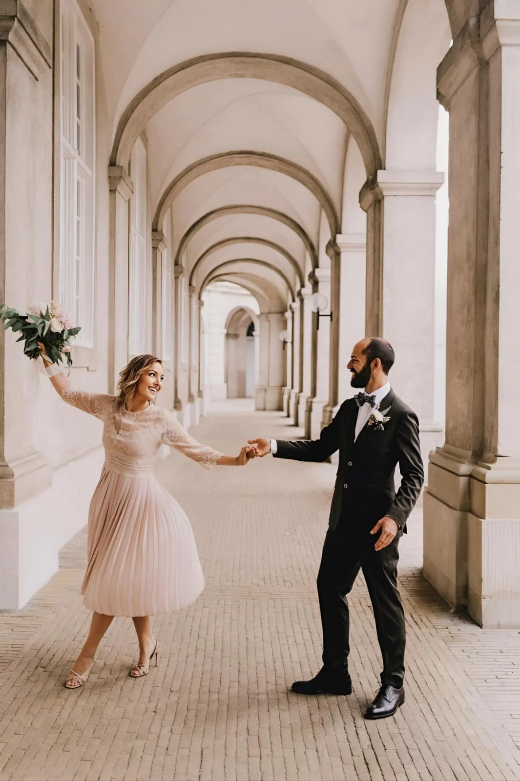 Ein verliebtes Paar tanzt in einer eleganten, porzellanfarbenen Bogenhalle. Die Frau trägt ein langes, rosafarbenes Kleid mit Falten am Rock, während der Mann einen schwarzen Anzug mit Fliege trägt.
