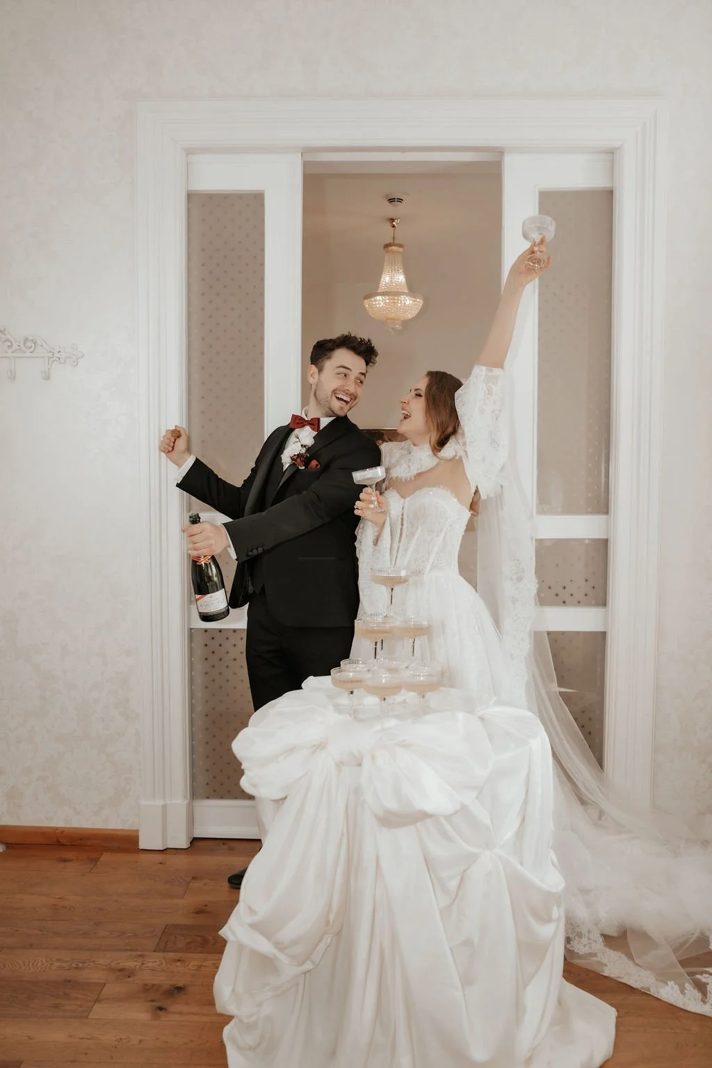 Ein Brautpaar beim Anstoßen bei ihrer Hochzeit, sie jubelt mit einem Glas, er hält eine Flasche Sekt, im Vordergrund ein Tisch mit einem Champagner-Turm, im Hintergrund eine elegante Innenraum mit Kronleuchtern.