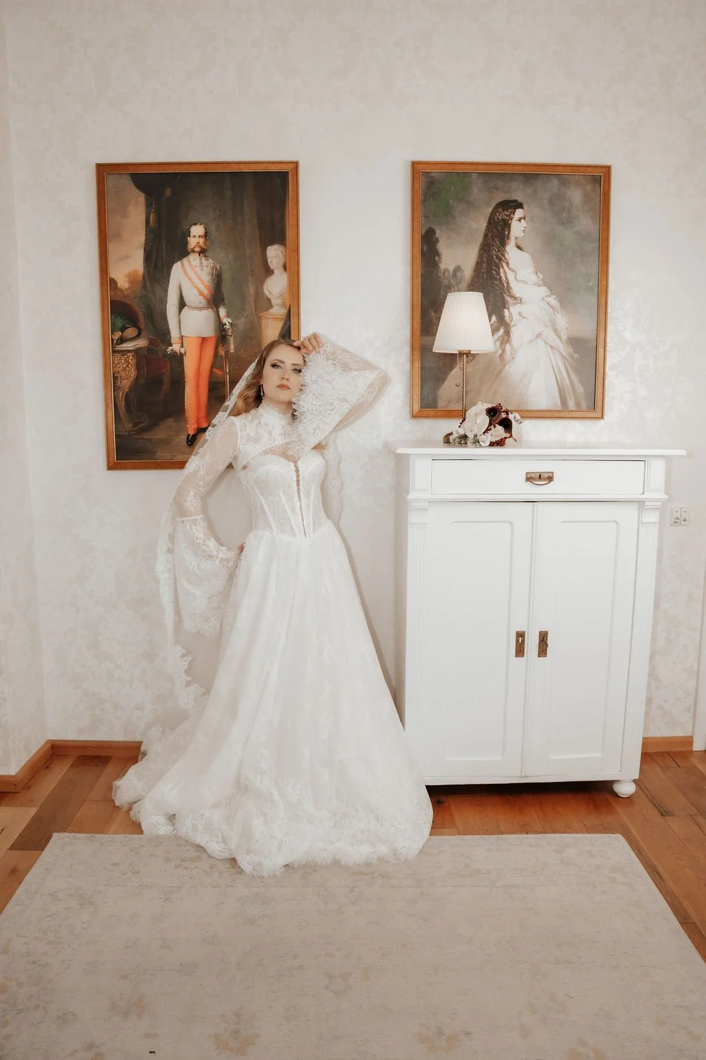 Junge Frau in weißem Brautkleid steht vor einer weißen Kommode mit einem Blumenstrauß und Tischlampe, hinter ihr Bilder von historischen Persönlichkeiten an der Wand.