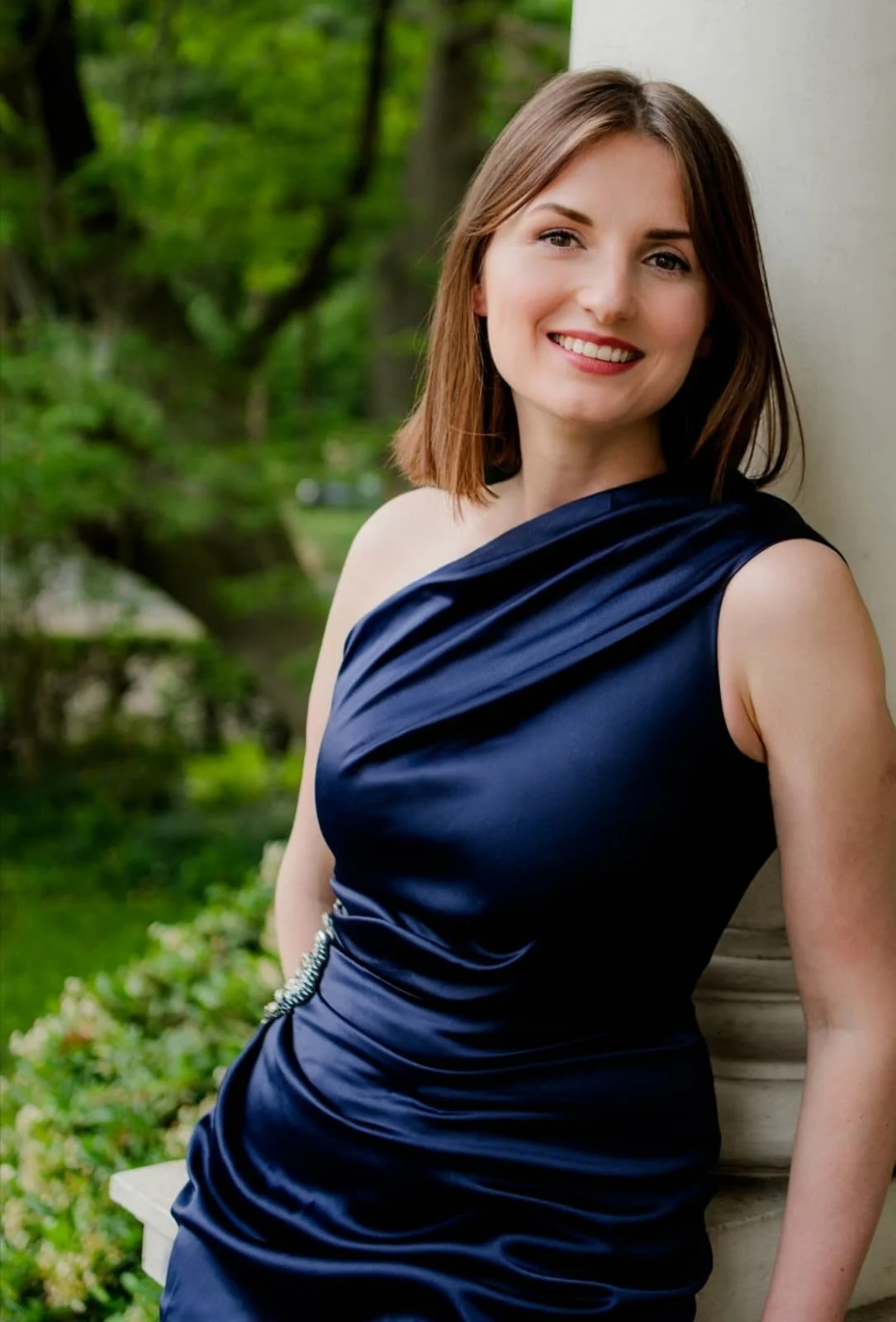 Junge Frau in dunklem blauen Kleid, lächelnd, steht im Grünen neben einer weißen Säule.