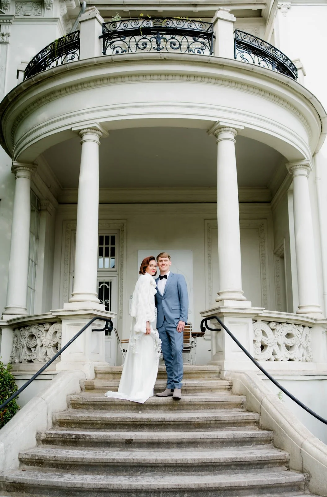 Ein Brautpaar steht auf den Stufen eines weißen, eleganten Hauses, das neoklassizistische Architektur mit Säulen, Balkon und detaillierten Verzierungen aufweist. Die Frau trägt ein weißes Hochzeitskleid mit Pelzmantel, der Mann einen blauen Anzug mit