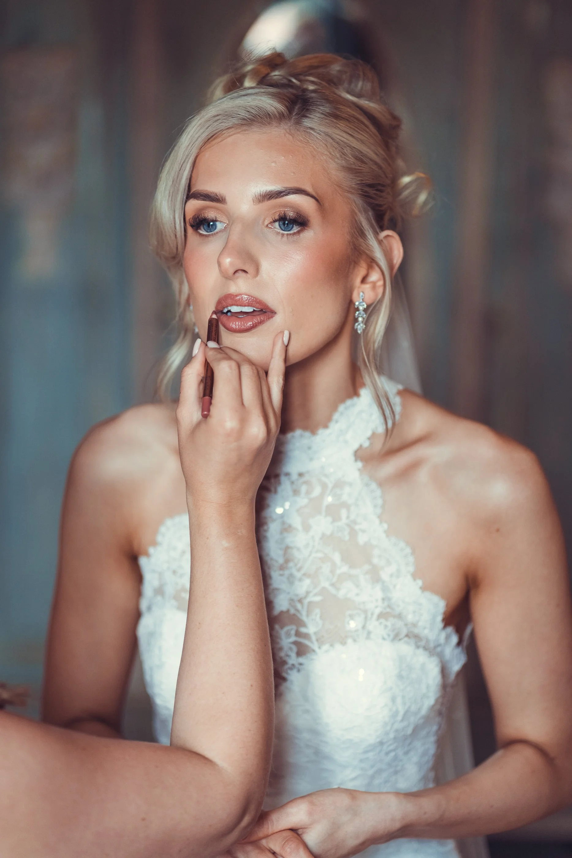 A bride with blonde hair and blue eyes is getting her makeup done, with someone applying lipstick to her lips. She is wearing a lace wedding dress and earrings.