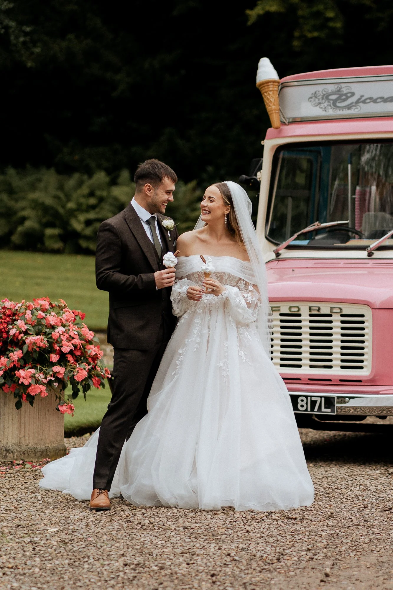 A bride and groom in wedding attire standing outdoors near a pink ice cream truck, both smiling and holding ice cream cones.