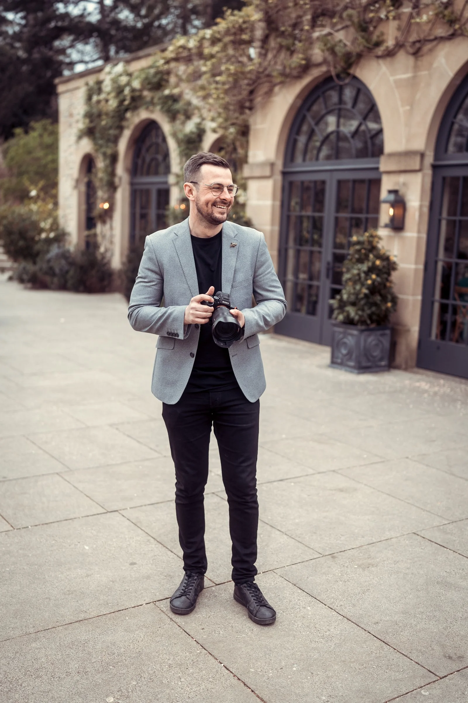 A man smiling, holding a camera, standing outdoors in front of a building with large arched windows.