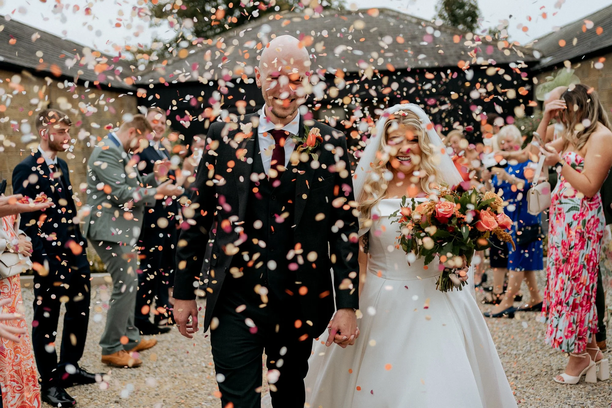 A newlywed couple walking hand in hand through confetti during their wedding celebration, with guests surrounding them and throwing confetti in the air.