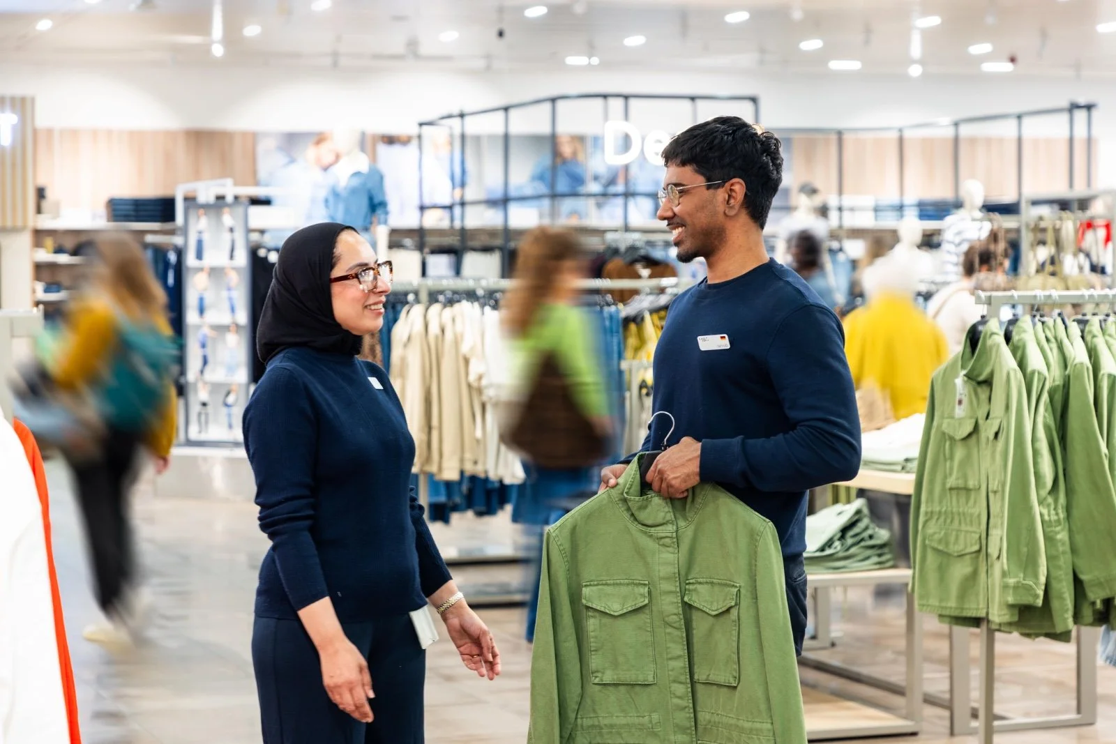 A man shopping for a green shirt in a clothing store, talking to a female employee with a hijab.