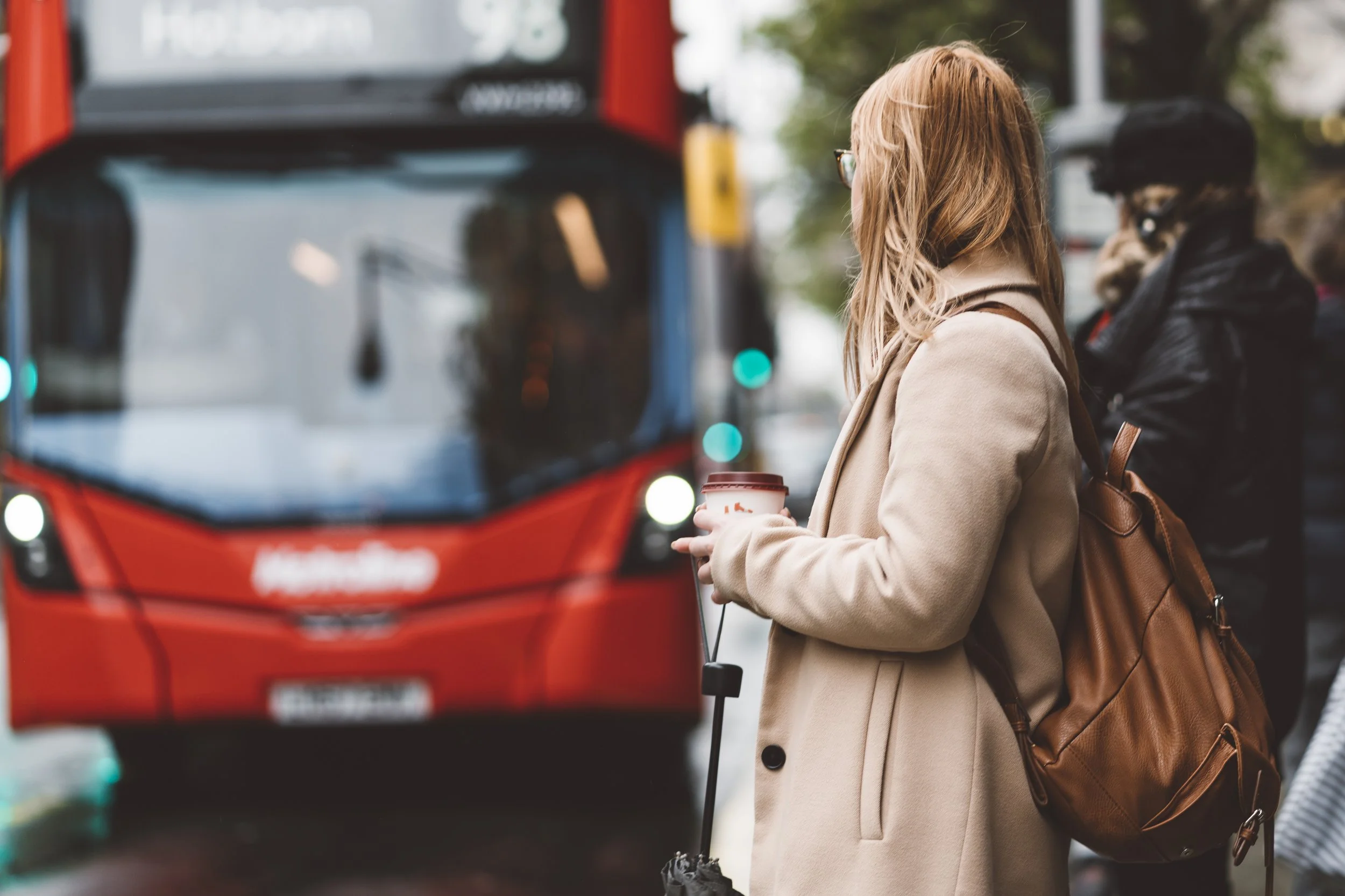 A woman with glasses and a brown backpack waiting at a bus stop, holding a coffee cup, with a red bus approaching.