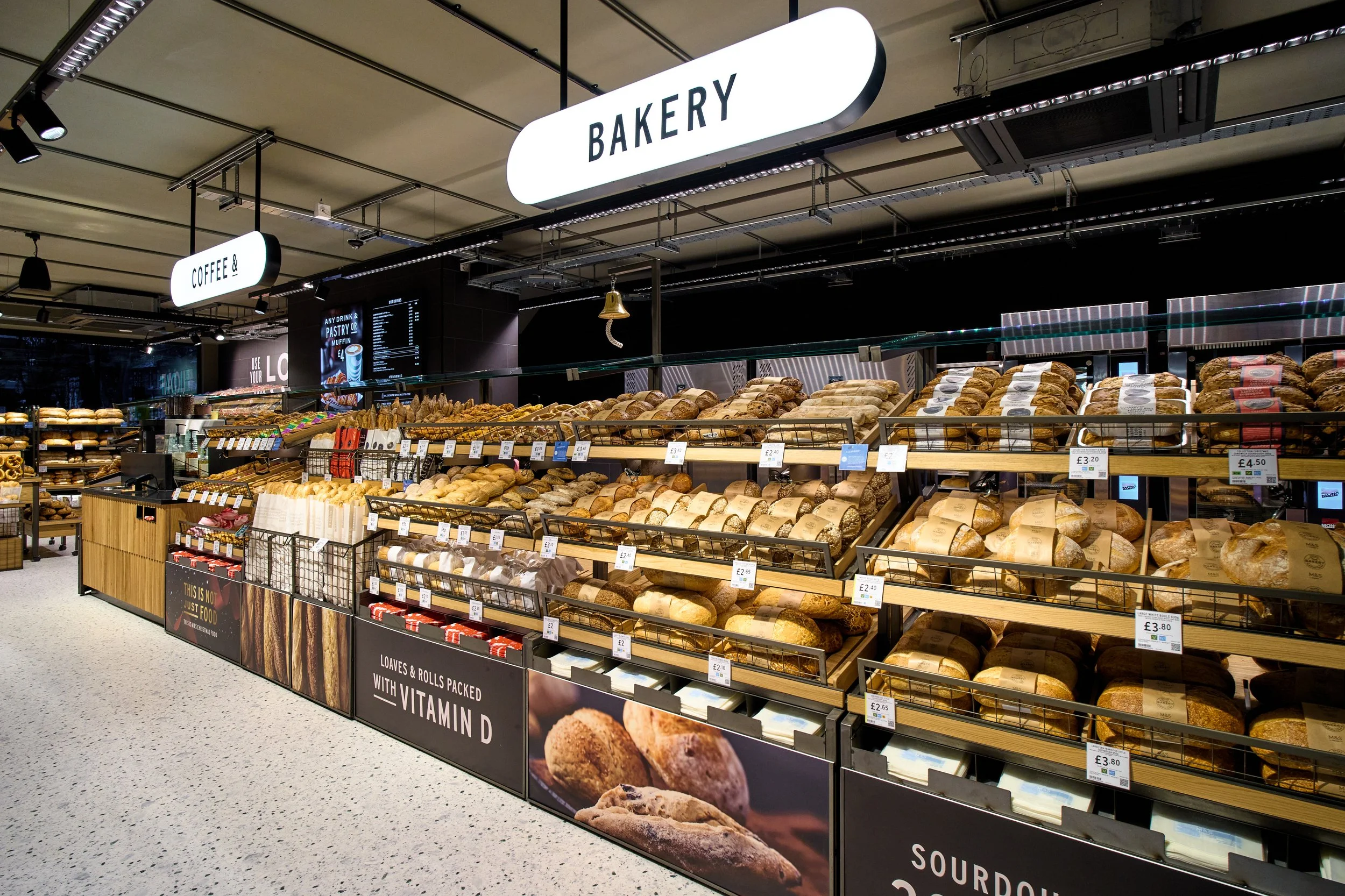 Inside a bakery section of a grocery store with shelves of bread and baked goods, illuminated signs reading 'BAKERY' and 'COFFEE &' are hanging from the ceiling.
