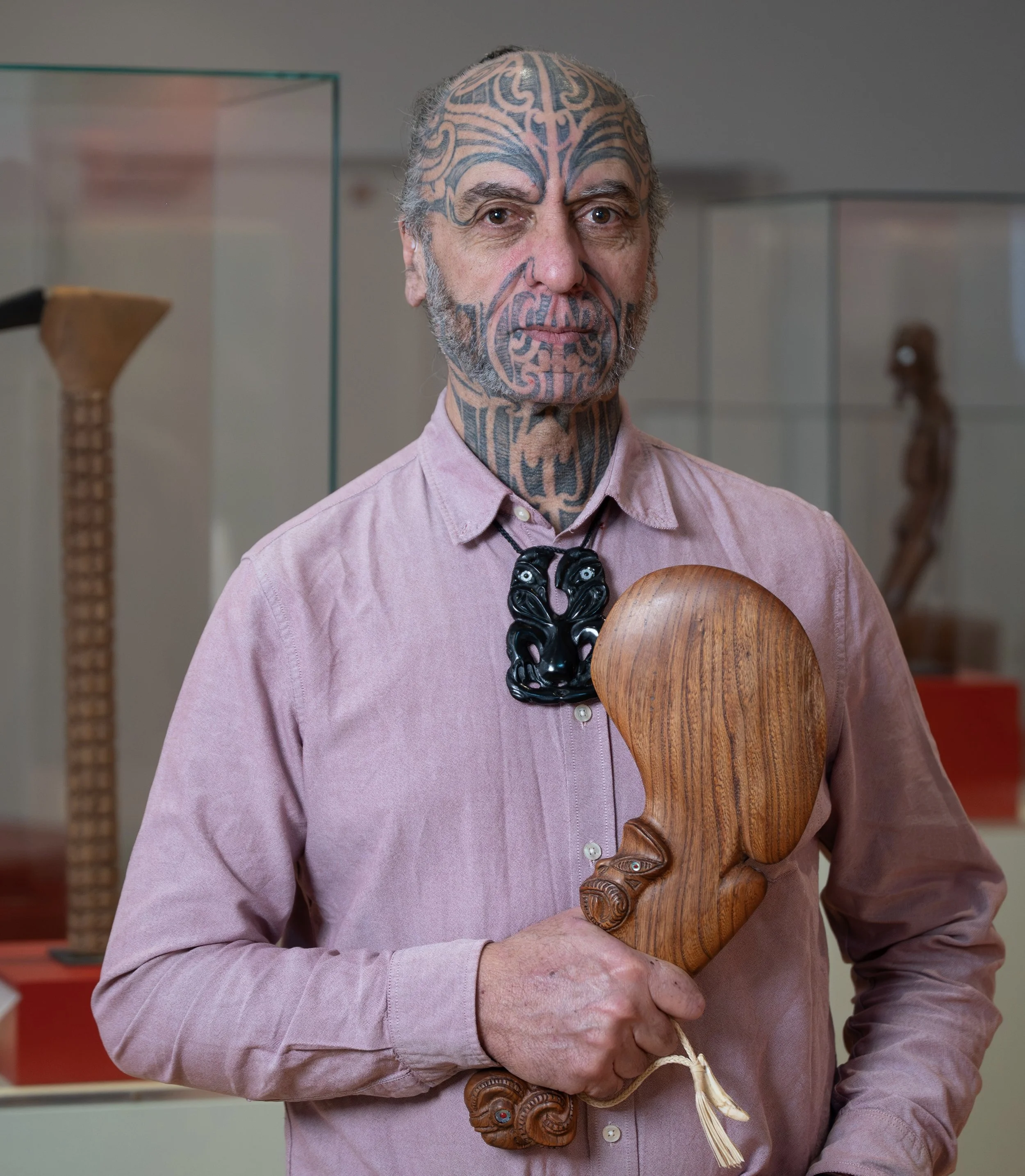 A man with extensive tattoos on his face and neck, wearing a pink button-up shirt and a black carved necklace, is holding a large wooden carved figure resembling a face. He stands in a display area with glass cases and other carvings visible in the background.