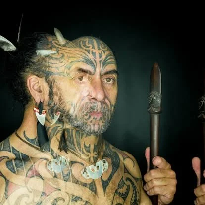 A person with extensive tribal tattoos and facial tattoos, holding a traditional spear or staff, with feathers in their hair.