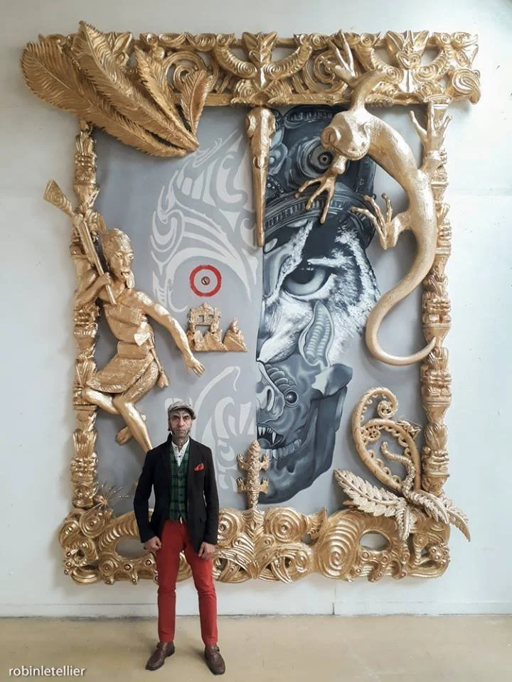 A man in a black blazer, red pants, and brown shoes stands in front of an elaborate, ornate gold frame with various mythical and fantastical sculptures. Inside the frame, there is a large painting of a tiger's face, artistically stylized with additional abstract designs, and a person wielding a spear. The frame features a dragon head, a snake, a feather, and other intricate decorative elements.