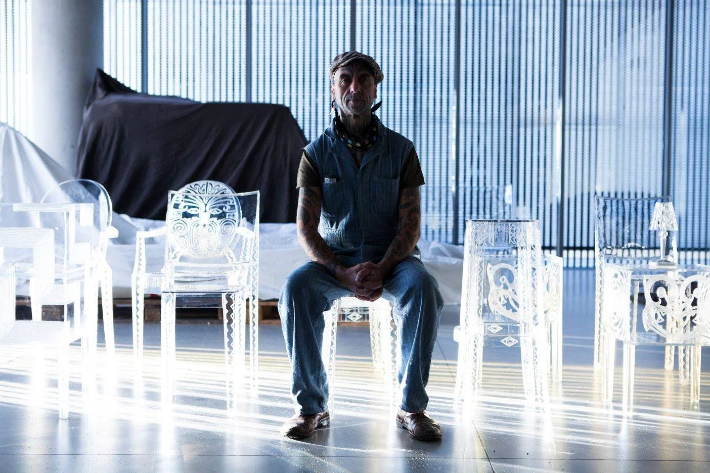 A man with tattoos and a beard sitting in a brightly lit modern space surrounded by clear, ornate plastic chairs with intricate designs, with large windows with vertical blinds in the background.