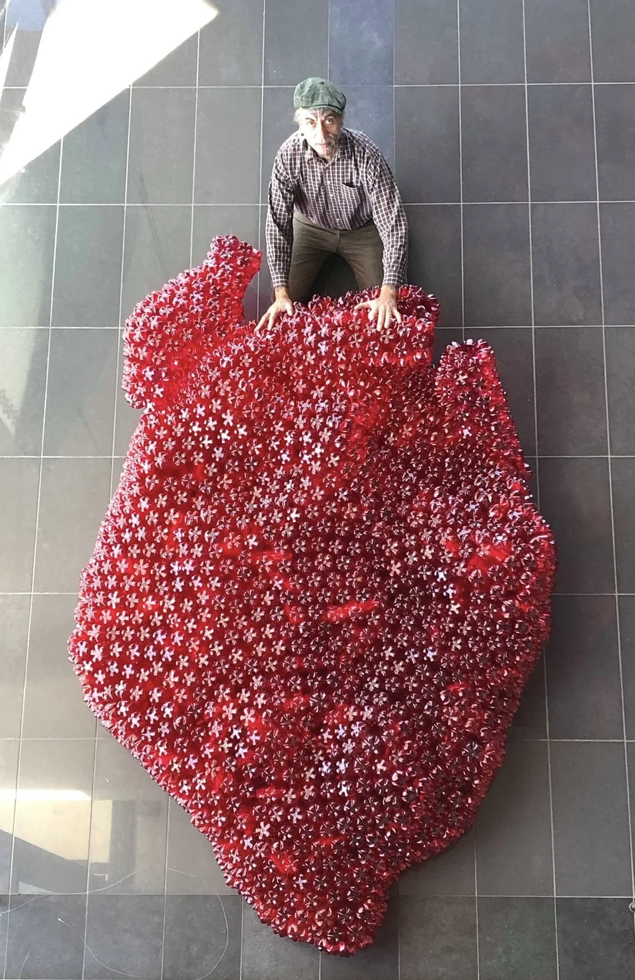 A man in a plaid shirt and a cap kneeling on a red sculptural art piece resembling an abstract flower or plant, on a gray tiled floor.