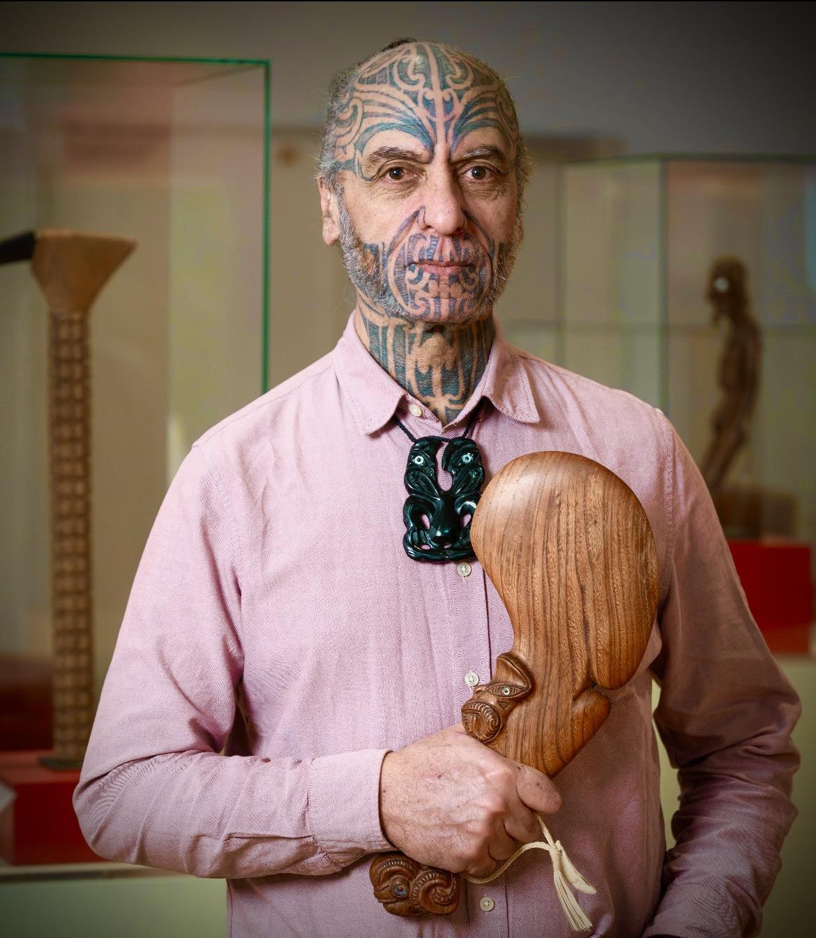 A man with extensive facial and neck tattoos holding a wooden carving of an abstract figure, standing in what appears to be a museum or gallery with display cases in the background.