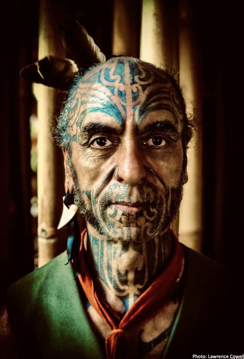 A man with extensive facial and scalp tattoos, wearing a green garment and a red scarf, has animal-like ears attached to his head, with a bamboo background behind him.