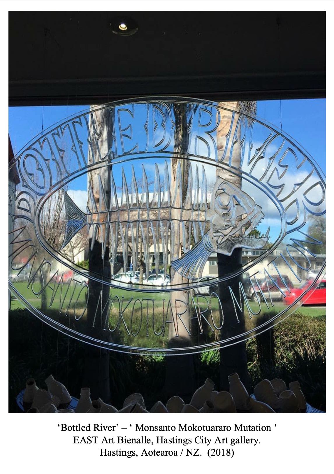 Reflective glass window with the words 'Bottled River' and 'Monsanto Mokotuaro Mutation' etched in it, showing outdoor trees, a blue sky, and cars in the background.