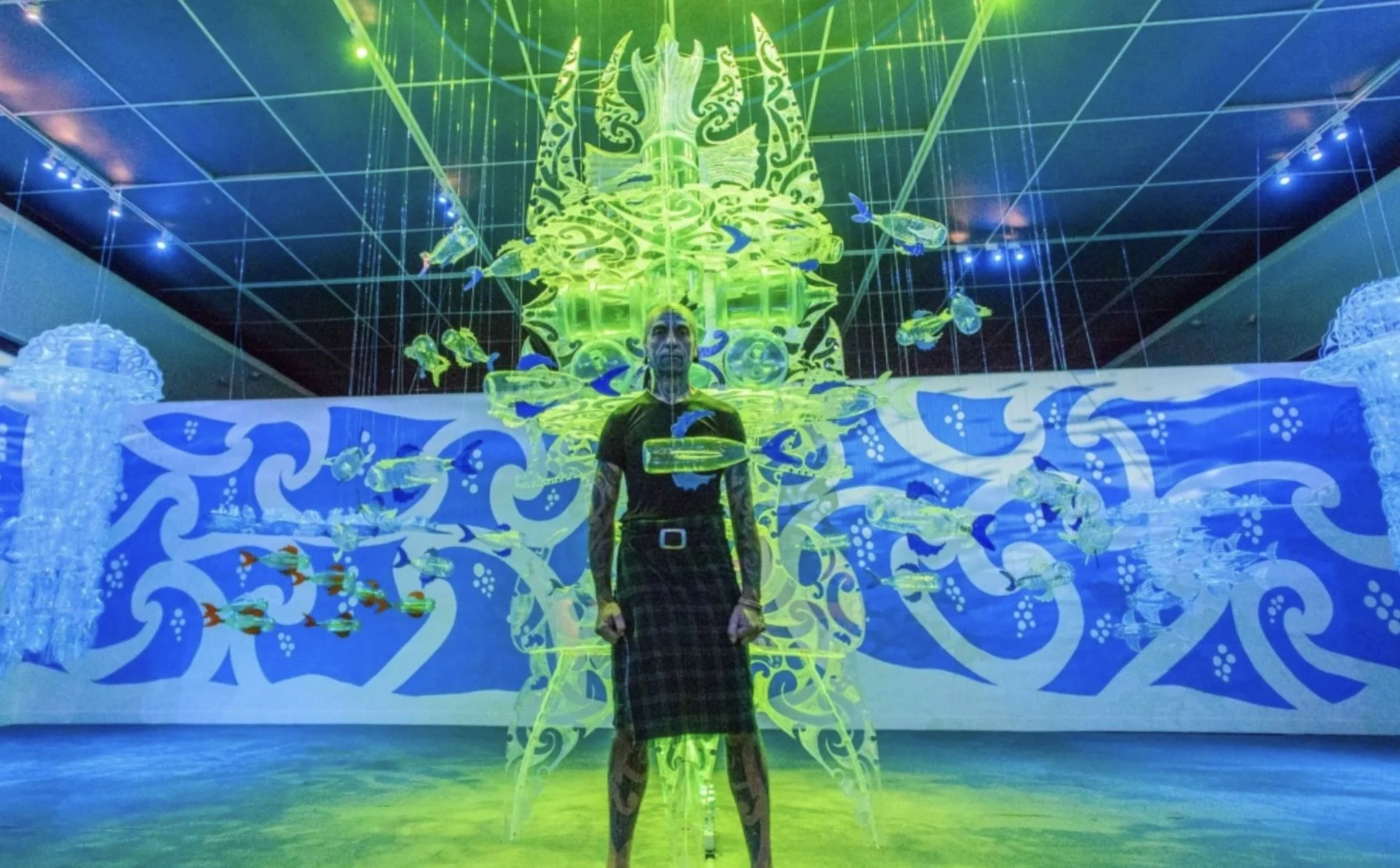 Person standing in front of an illuminated, ornate, green and yellow sculpture of a bird, with fish swimming around in an indoor setting with vibrant blue and green lighting.