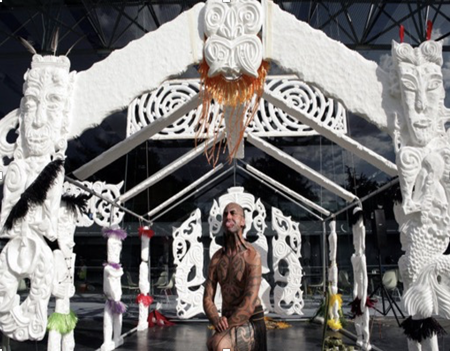 A man with extensive tattoos poses in front of a large, intricate white Maori-style carved wooden structure decorated with colorful feathers and adorned with a tiki face in an outdoor setting.