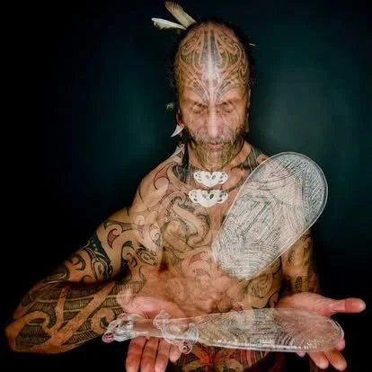 Tattooed man with traditional face paint holding a clear paddle and a translucent feather fan.