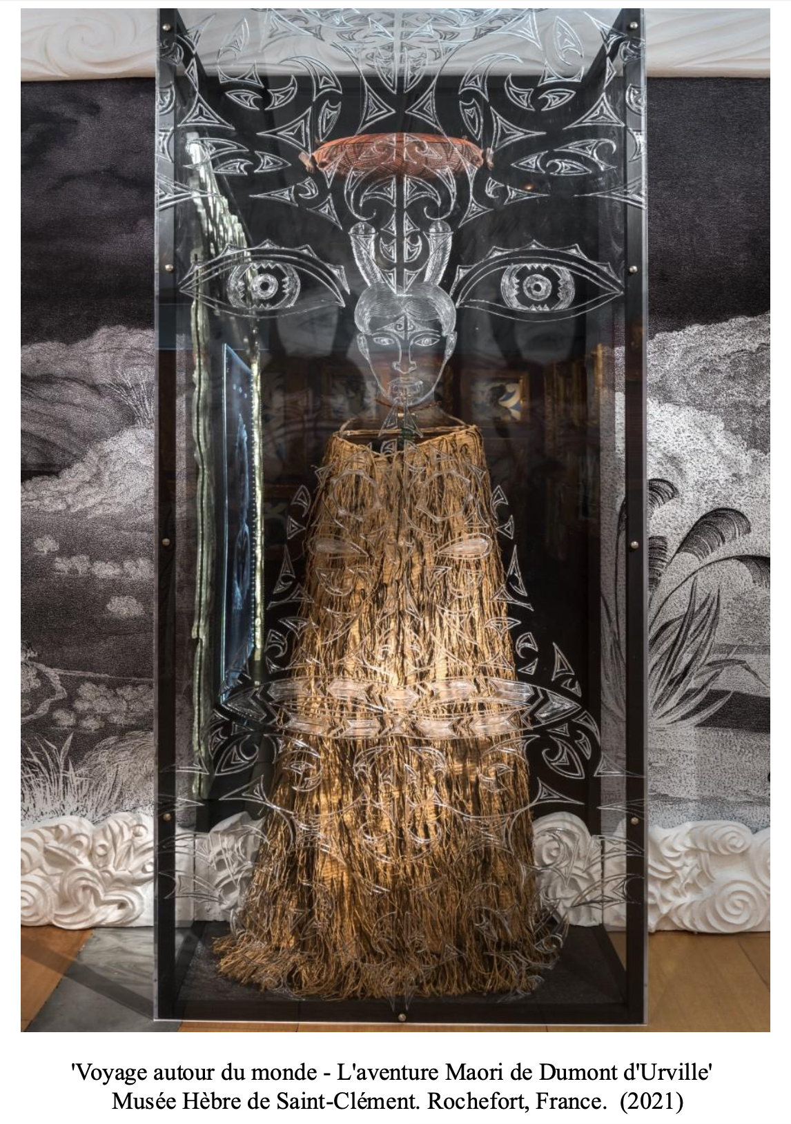 An art installation of a Maori doll with a woven straw skirt in front of a glass panel with etched facial features and eyes, set inside a display case.