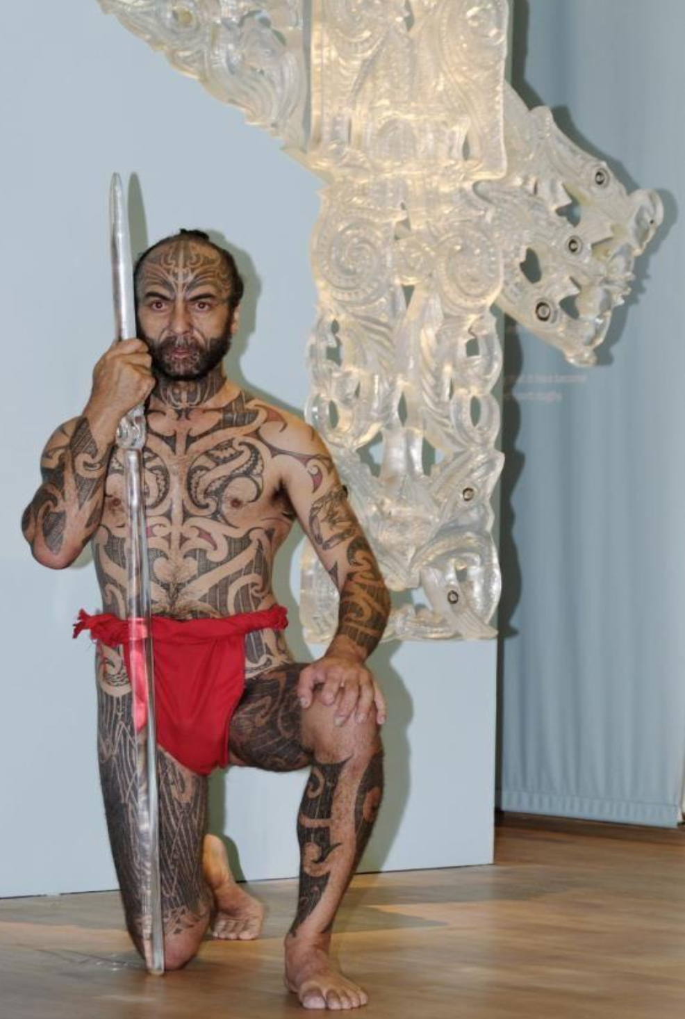 Man with traditional Polynesian tattoos kneeling indoors, holding a spear, wearing red cloth, next to a large Polynesian-style carved wooden sculpture.