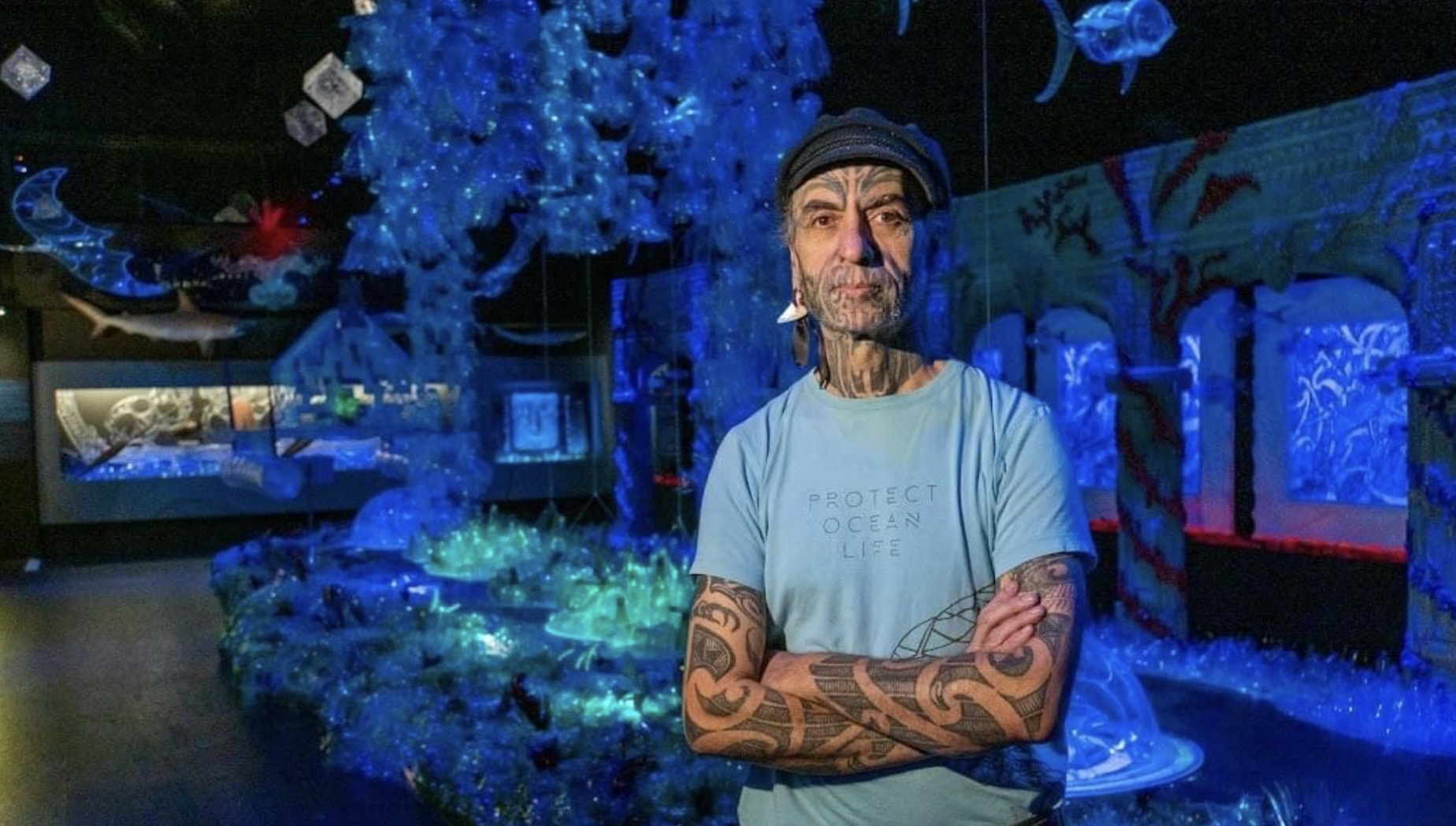 A man with tattoos and a black cap poses with arms crossed inside an aquarium exhibit filled with blue and green lighting and various sea creatures, including fish and jellyfish.
