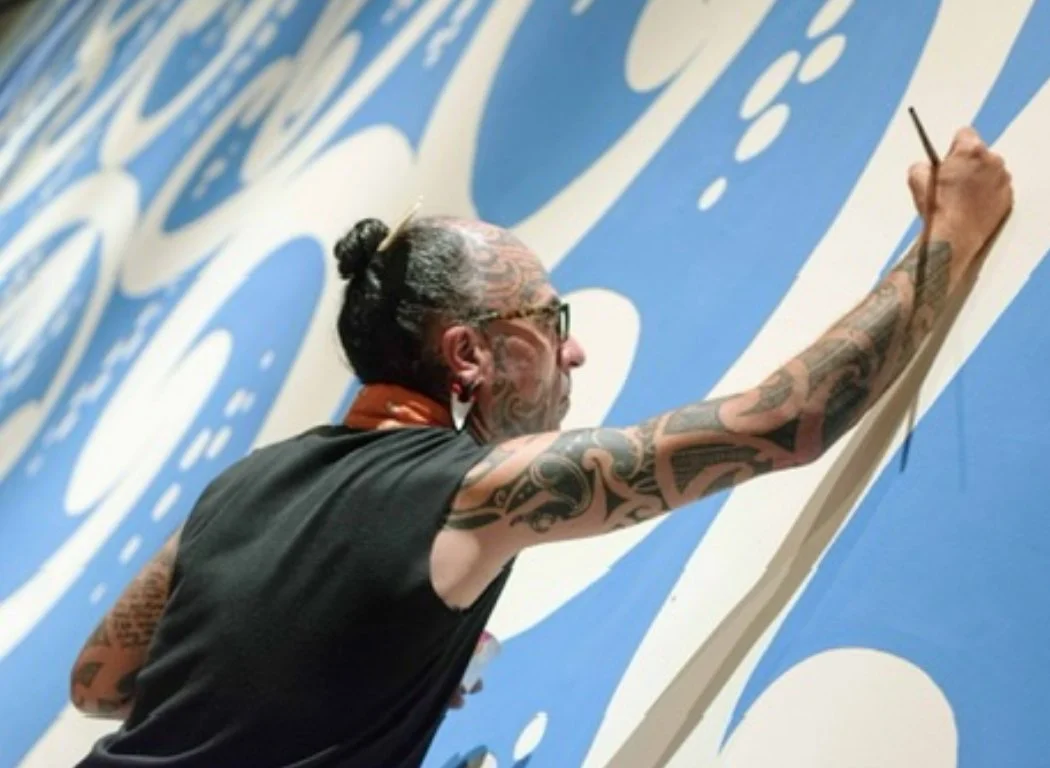 A tattooed man with glasses and a bun hairstyle painting a large mural with a blue background and white design.