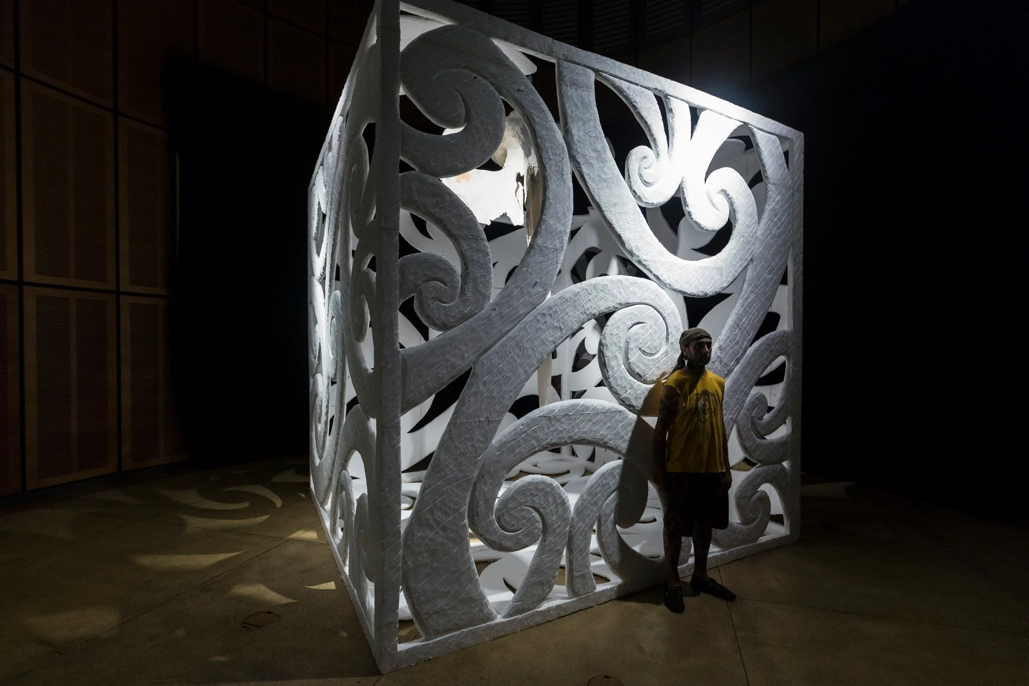 A person wearing a yellow shirt and shorts stands in front of a large, white, ornate, three-dimensional decorative panel with swirling patterns. The panel is illuminated from behind, casting shadows on the floor. The person is standing inside a dimly lit room with wooden panels on the wall.