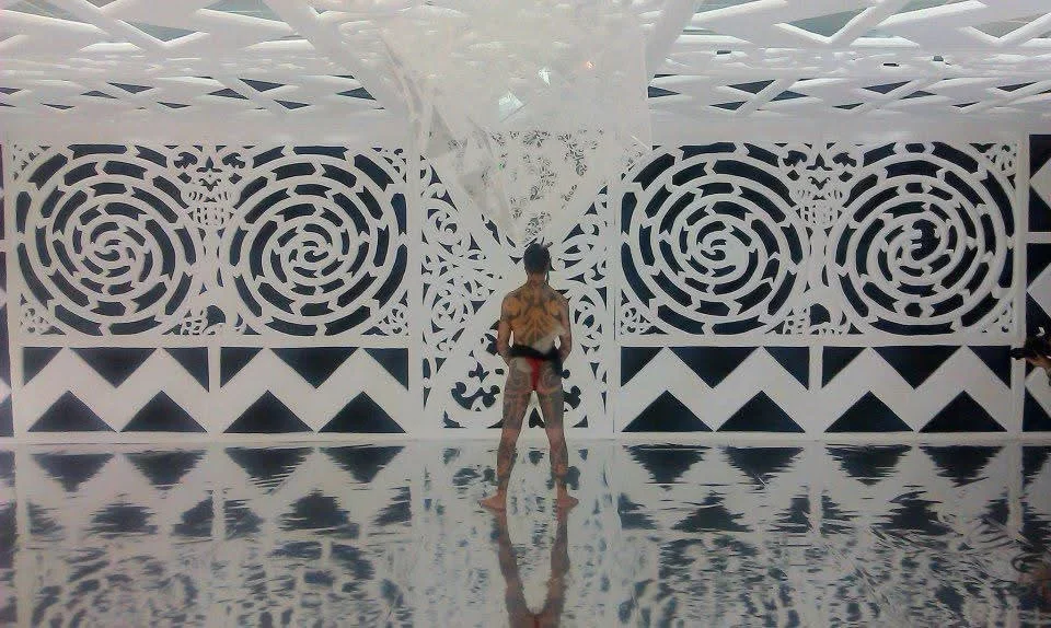 A shirtless man with body paint standing on a reflective floor in front of an ornate white wall with circular and swirling patterns.