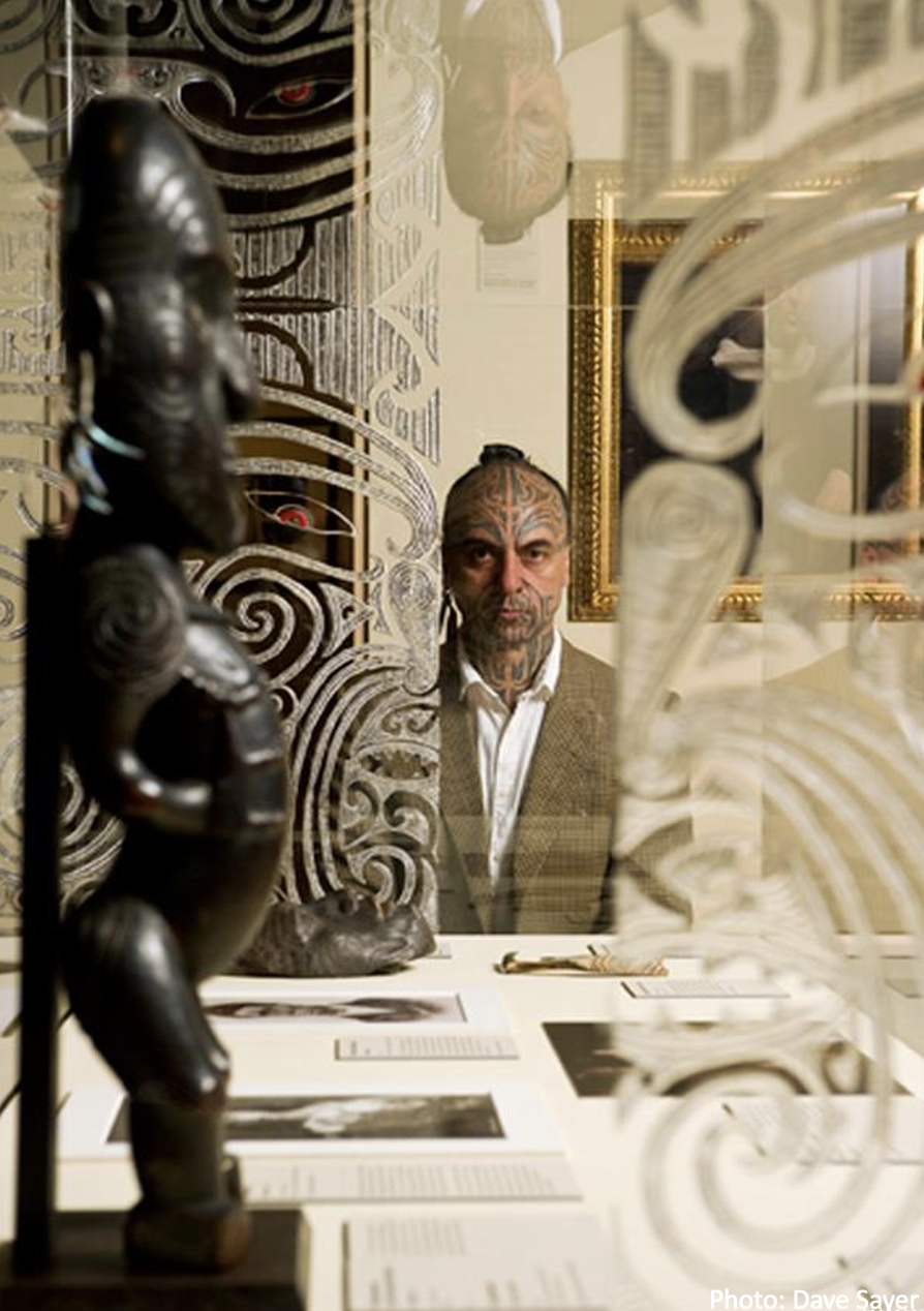 A man with tattoos on his face and head, dressed in a suit jacket and white shirt, standing behind a display case with art pieces and photographs, looking through decorative glass.