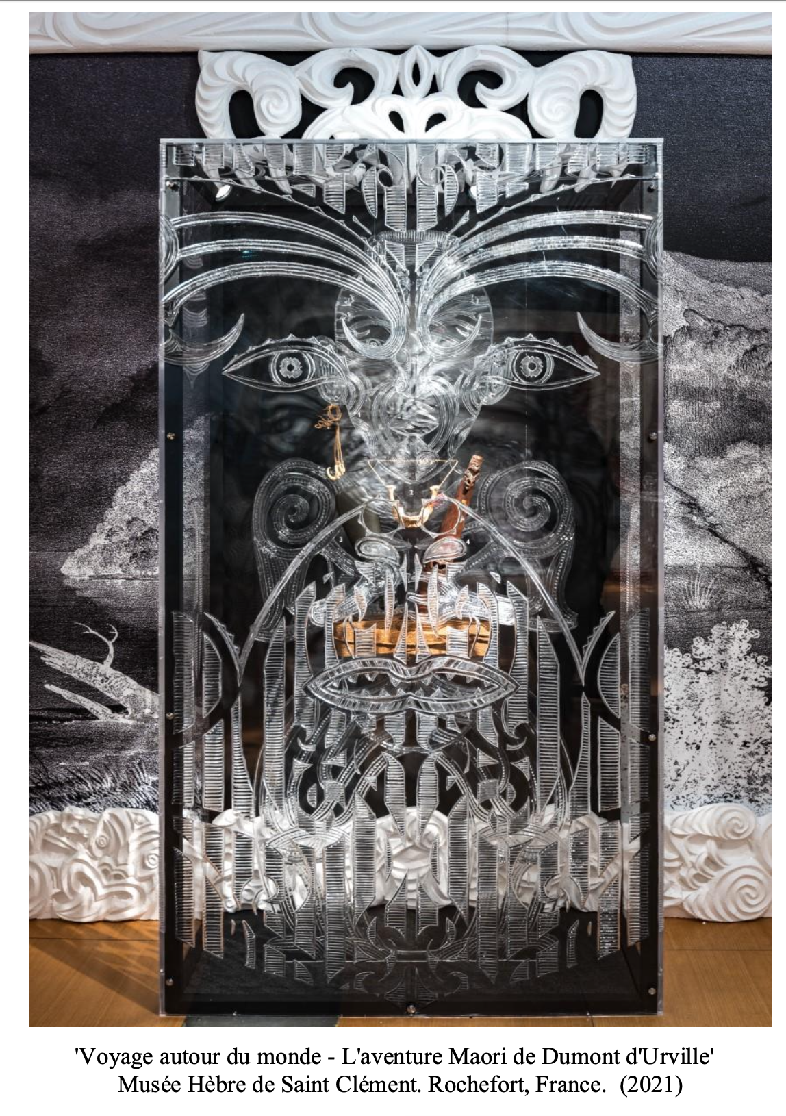 A carved glass artwork of a Maori face with detailed facial features, horns, and intricate patterns, displayed in front of a black and white background.