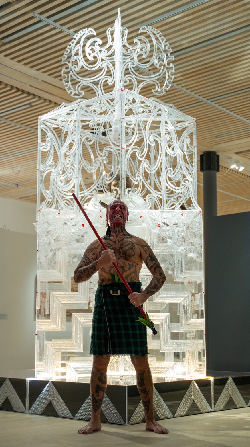 Tattooed man with traditional Maori tattoos, wearing a kilt, holding a spear, standing in front of a decorative Polynesian sculpture at an indoor art exhibit.