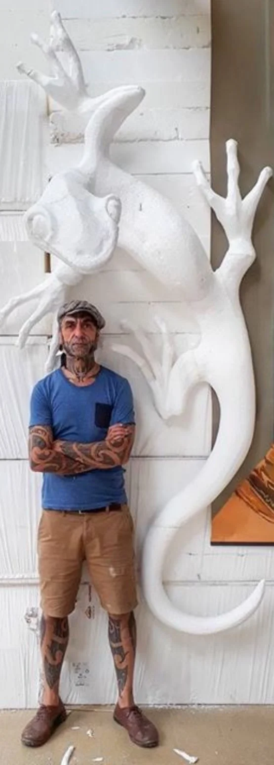A man with tattoos, wearing a blue t-shirt, beige shorts, and a gray cap, stands with arms crossed in front of a large white sculpture of a seahorse.