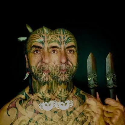 A person with extensive face and body tattoos holding large knives, with a dark background.