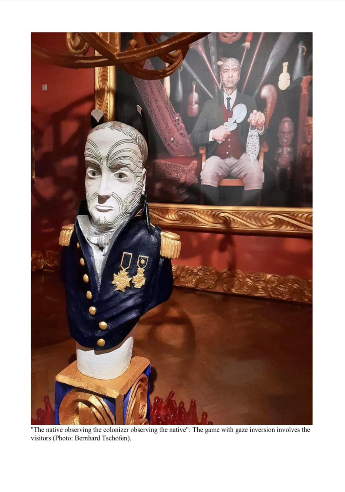 A detailed sculpture of a colonial officer with a serious expression, dressed in a navy uniform with medals, in front of a framed historical artwork depicting a man with a goat's head in a formal setting surrounded by other colonial and indigenous artifacts.