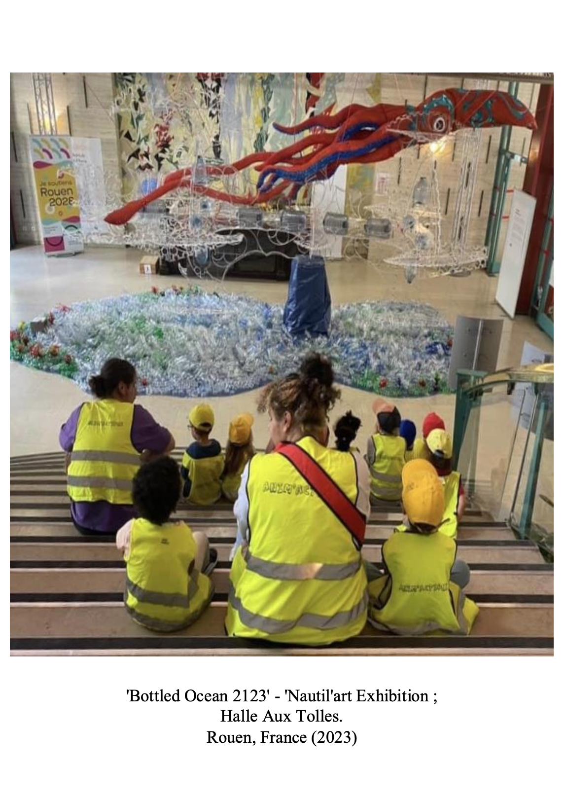 Children and adults wearing yellow safety vests sitting on stairs and observing an art installation made of plastic bottles resembling an ocean scene with a large octopus sculpture hanging overhead.