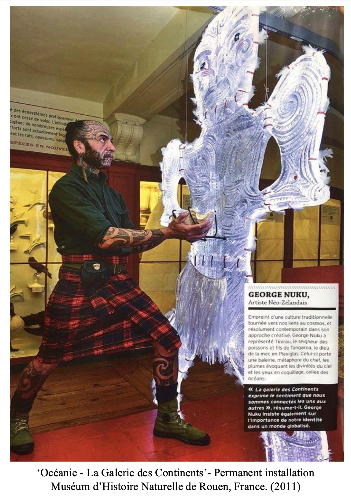 An artist wearing traditional Scottish attire and with tattoos on his arms is working on a large illuminated art piece that is shaped like a human figure. The setting appears to be a museum exhibit, with display cases and informational panels in the background.