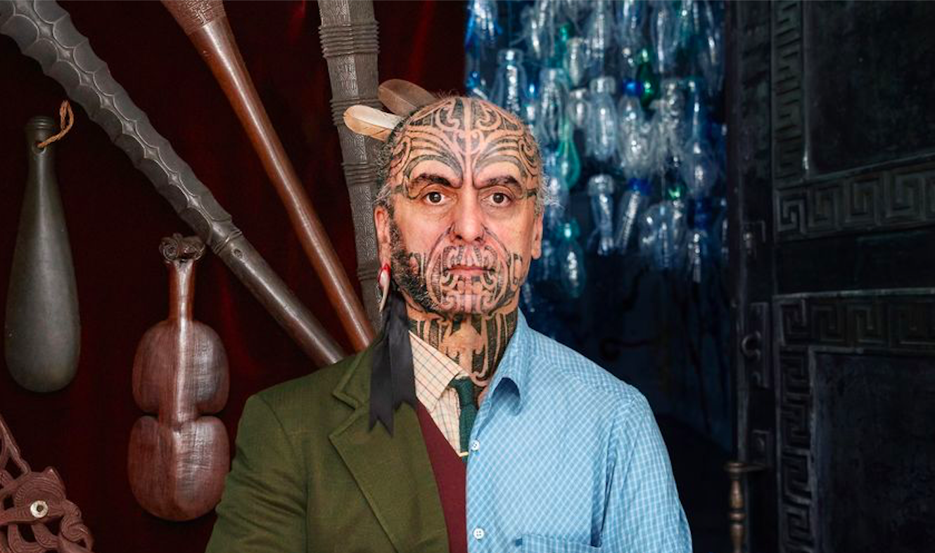 A man with extensive tattoos on his face, neck, and ear, dressed in a blue checkered shirt and a green jacket, standing in front of wooden and glass art pieces on a dark background.