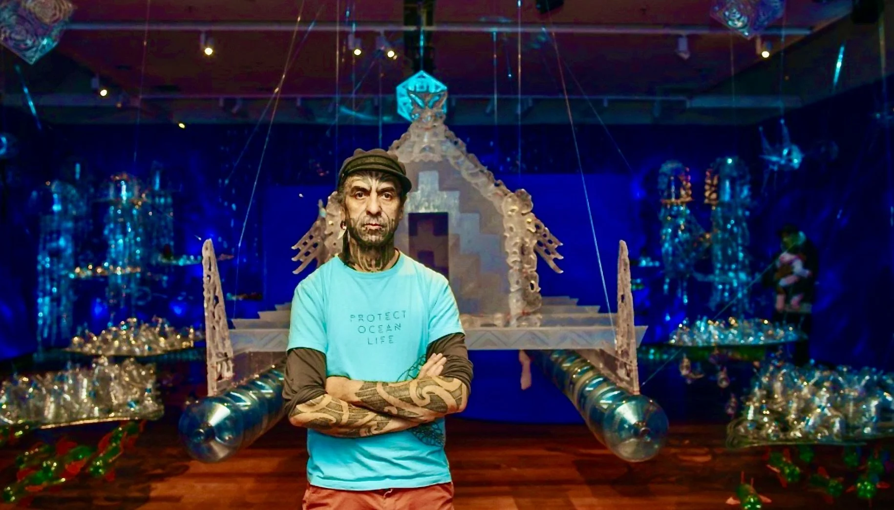 A man with tattoos and a beard, wearing a blue t-shirt and red shorts, standing with arms crossed in an indoor exhibit with various sculptures and art installations.