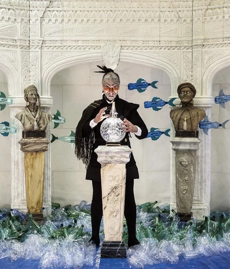 A man dressed in dark clothing with elaborate face makeup, holding a glowing orb, stands behind a marble pedestal in a decorative room with white carved walls. There are two classical busts on pedestals, with plastic bottles scattered on the floor, and colorful fish-shaped objects floating around.