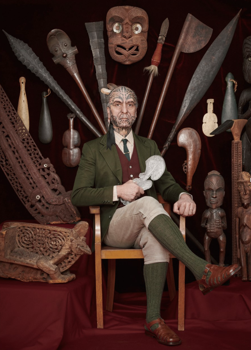 A man dressed in vintage clothing sits on a wooden chair surrounded by traditional African tribal masks and carvings. He holds a carved object and is set against a dark red backdrop.