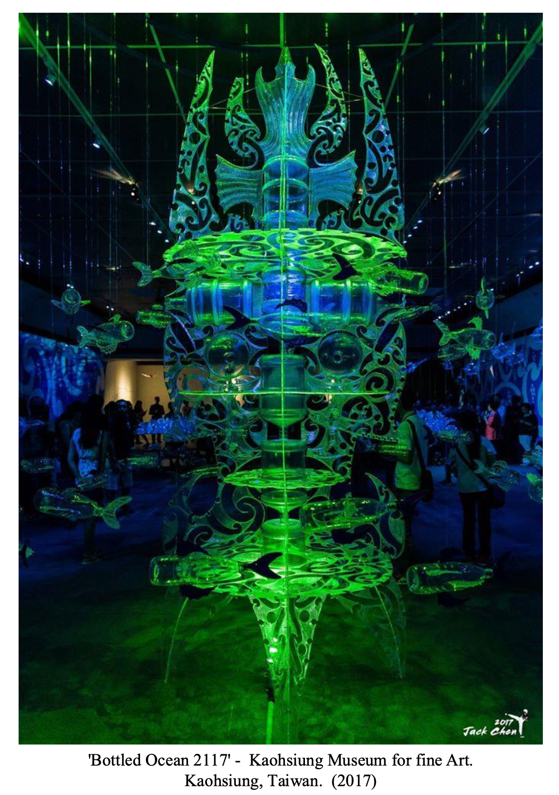 A glowing, intricate art installation called 'Bottled Ocean 2117' at the Kaohsiung Museum for Fine Art in Taiwan, 2017. It features a large vertical structure with detailed swirling patterns resembling ocean life, glowing in green and blue lights, surrounded by suspended fish shapes. Visitors are seen observing the piece in a dimly lit room.