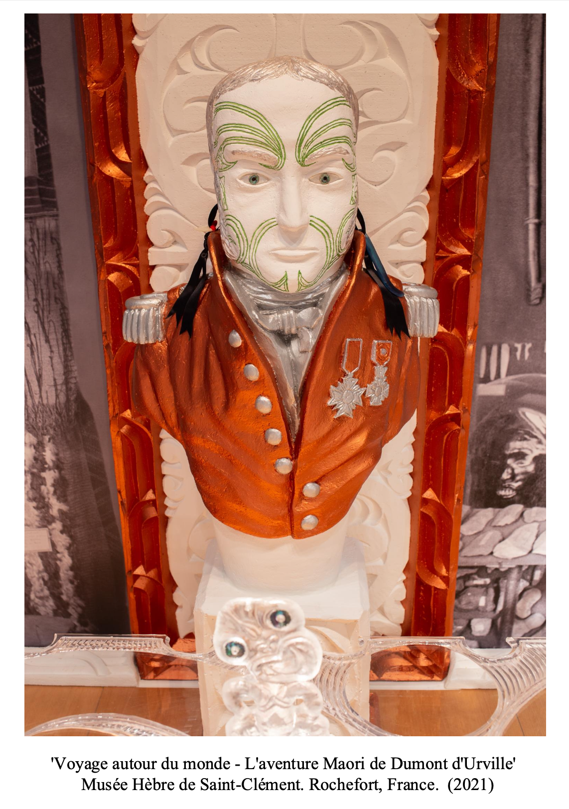 A carved wooden bust of a man dressed in a military uniform with medals, with green tribal face paint, set against a decorative background. The display is part of an exhibit at the Musée Hébre de Saint-Clément in Rochefort, France.