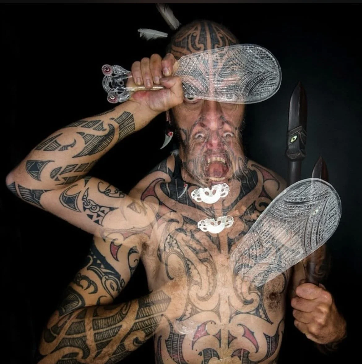 A person with extensive tattoos covering their arms and chest, holding carved wooden knives in each hand, and holding ornate glass blades in front of their face. Their facial expression is intense with an open mouth, possibly yelling or screaming, and their hair appears to have traditional Polynesian style markings.