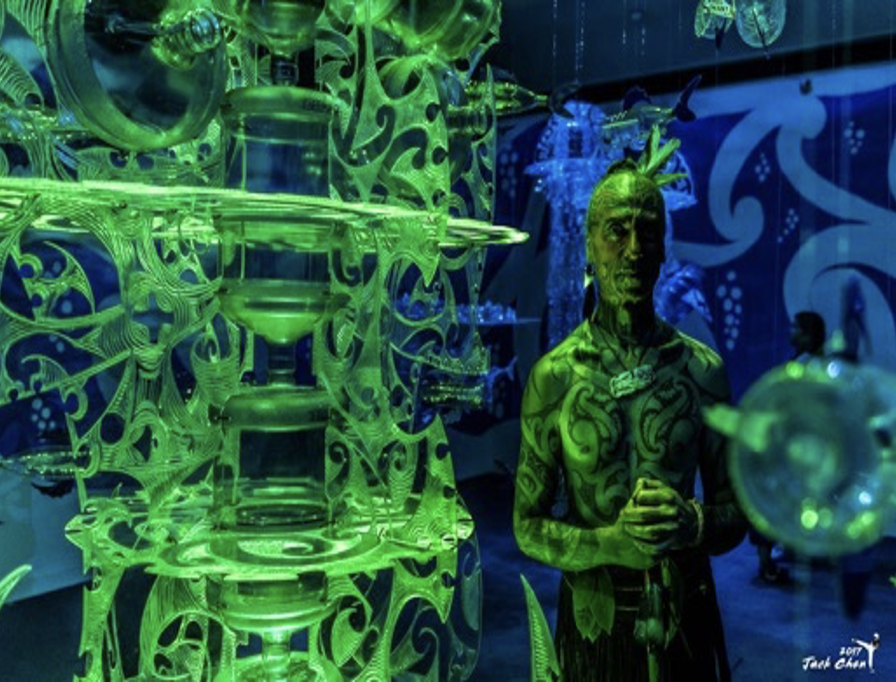 Person with body painted to resemble glowing green plant or alien figure standing in a room with illuminated green and blue patterned decorations, illuminated objects, and other art installations.