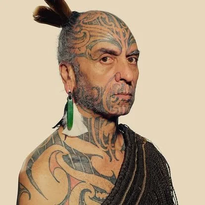 A person with extensive facial and body tattoos, wearing a green earring and a black shawl, with hair styled in a topknot.