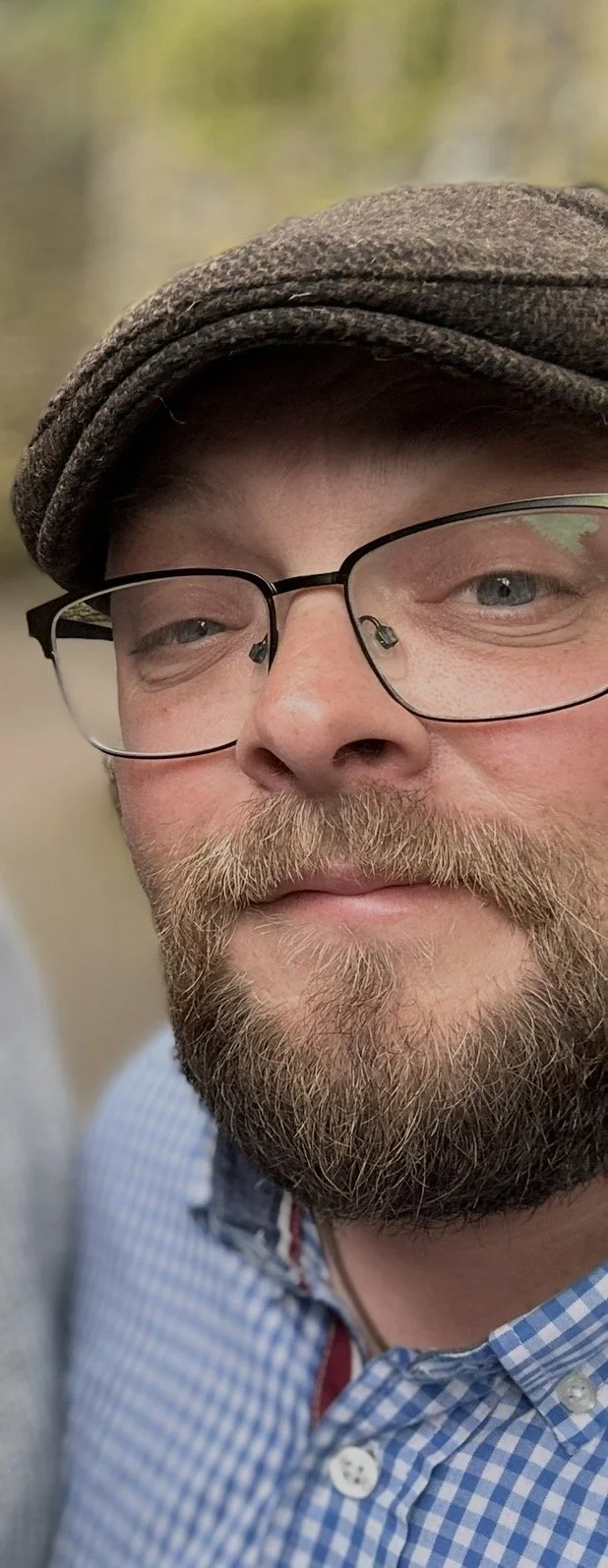 Close-up of a man with glasses, a beard, and a mustache, wearing a flat cap and a blue checkered shirt, smiling outdoors.