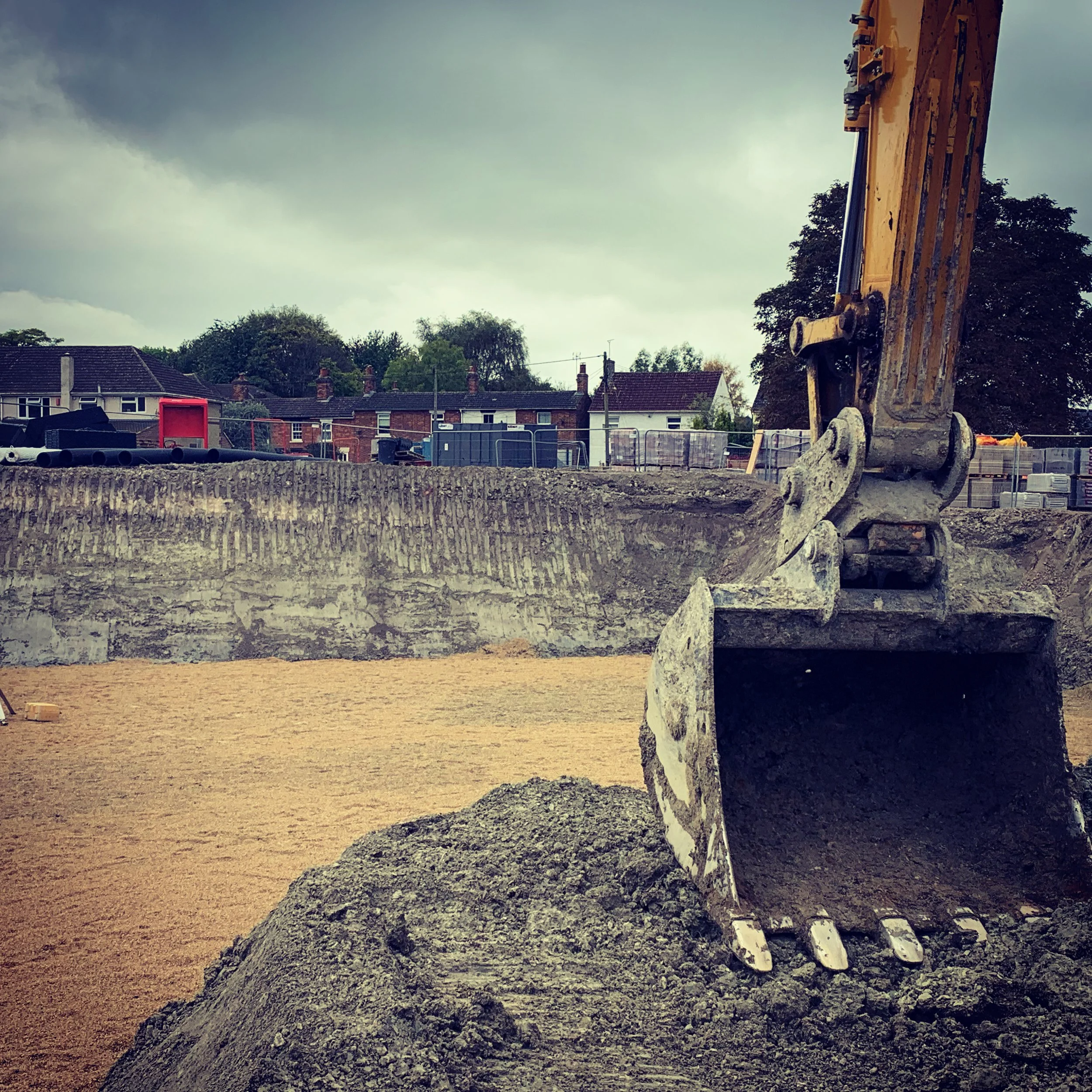 Close-up of an excavator bucket on a construction site, with dirt and a partially excavated ground in the background, along with houses and trees under an overcast sky.